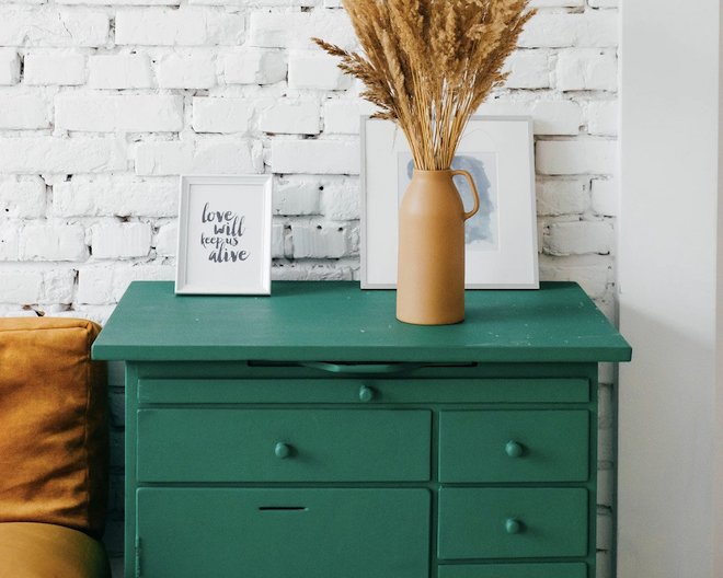 A dark green chest of drawers with three items on top of it: a decorative vase of wheat, a small picture frame that says 'love will keep us alive' and a painting.