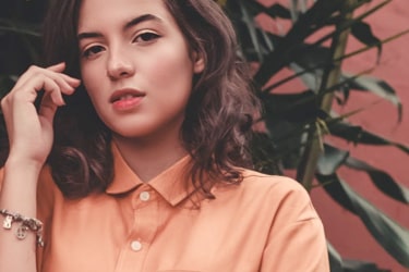 A model with brown eyes and shoulder-length brown hair. She is wearing an orange blouse with buttons and has a bracelet on her right wrist.