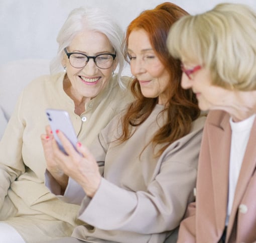 Three elderly women who are sitting together. They are in the middle of a video chat on a smartphone used by the one sitting in the middle.